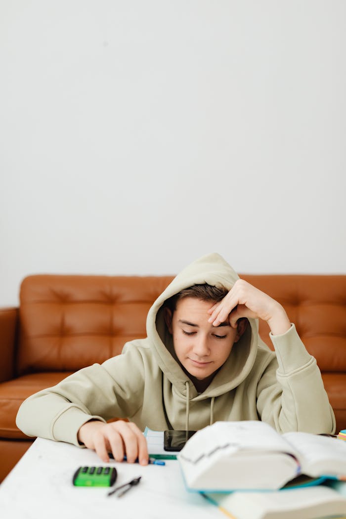 Teenager in a hoodie studying at home with books and calculator on the table.